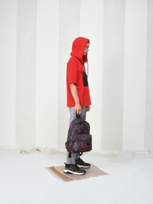 Male model wearing a red hoodie, grey trousers, brown trainers and a backpack in a print pattern by HUGO