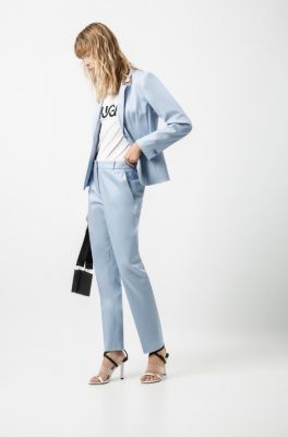 Female model wearing a blue jacket, white T-Shirt with HUGO logo, blue trousers, black bag and sandals by HUGO