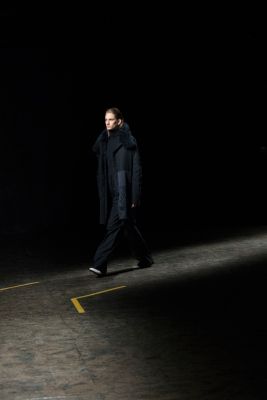 Woman on the runway wearing a full black look with black a black sweater, black trousers and a black down coat. All from the BOSS Womenswear Gallery Collection