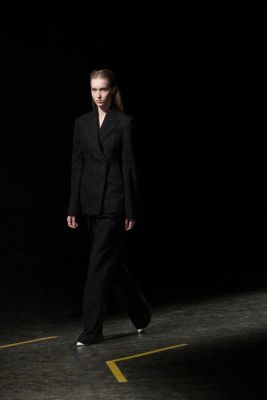 Woman on the runway wearing a full black look with black a black sweater, black trousers and a black down coat. All from the BOSS Womenswear Gallery Collection