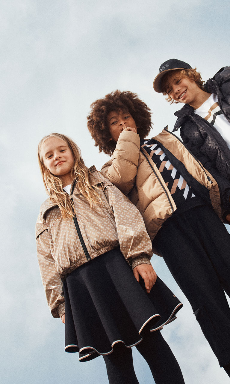 Girl in BOSS Kidswear camel jacket, white shirt and black skirt, boy in BOSS Kidswear camel outerwear jacket, white, camel and white sweater and black trousers, and boy in BOSS Kidswear black cap, black jacket and white shirt.  