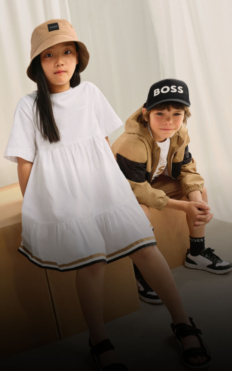 Girl in BOSS Kidswear camel jacket, white shirt and black skirt, boy in BOSS Kidswear camel outerwear jacket, white, camel and white sweater and black trousers, and boy in BOSS Kidswear black cap, black jacket and white shirt.  