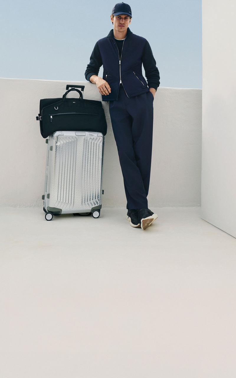male model in the new BOSS Performance tracksuit and a baseball cap all in navy blue leaning on a suitcase and a travelling bag.