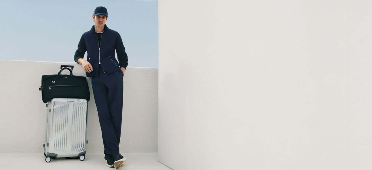 male model in the new BOSS Performance tracksuit and a baseball cap all in navy blue leaning on a suitcase and a travelling bag.
