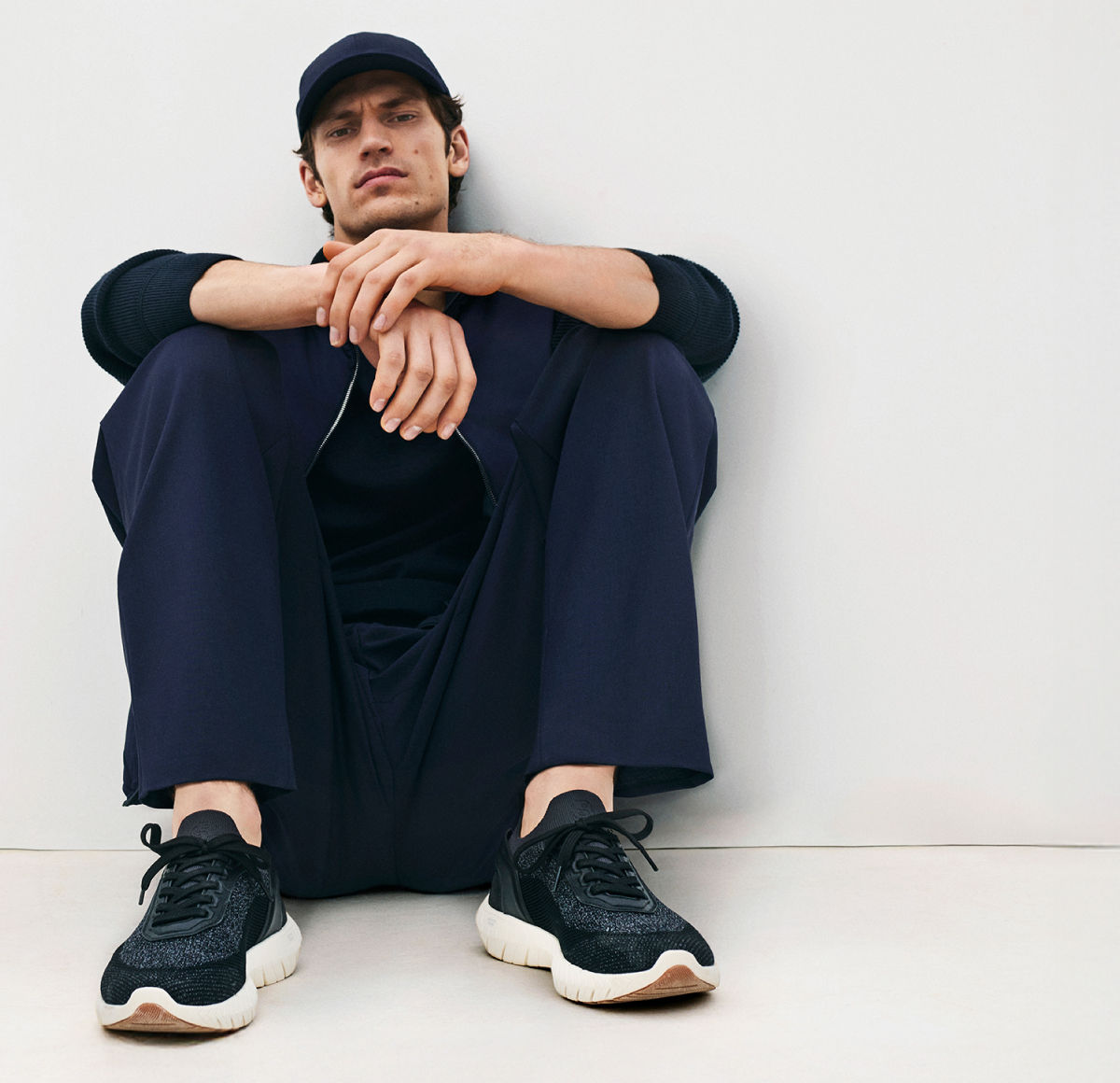 male model in the new BOSS Performance collection with a baseball cap and BOSS-Motion Sneakers all in navy blue, sitting in front of a whitewall